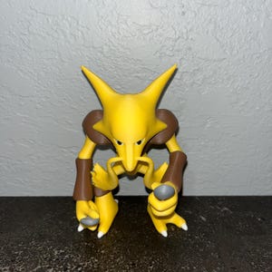 Könnte beinhalten: Gelb-braune Figur einer Kreatur mit spitzen Ohren, langer Nase und schnurrbartähnlichem Merkmal. Die Figur hat braune Arm- und Beinbänder sowie graue Akzente an Händen und Füßen. Die Figur steht auf einer dunklen Oberfläche.