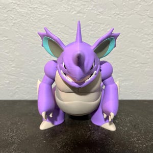 Puede incluir: Una figura morada de una criatura con vientre blanco, garras blancas y un cuerno. La criatura tiene orejas grandes con detalles turquesa. La figura está sobre una superficie oscura.