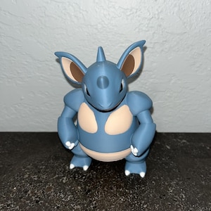 Puede incluir: Figura azul de un personaje de dibujos animados con orejas grandes y un cuerno. La figura tiene detalles en beige en las orejas y el pecho. El personaje está en cuclillas con los pies blancos.