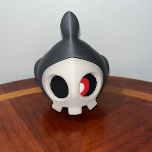 May include: A 3D-printed skull-shaped mask with a black hood and a white face. The eye sockets are black with a red crescent inside. The mask is sitting on a wooden surface.