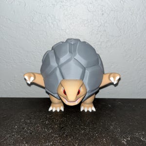 May include: A gray and tan figurine of a turtle-like creature with a segmented shell. The creature has tan arms, legs, and head, with red eyes and a smiling mouth. The figurine is standing on a dark surface.