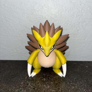 May include: A yellow and brown Sandslash figurine with white claws and a tan belly. The figure has a spiky back and a determined expression. The figurine is a collectible item.