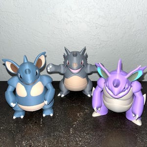 Puede incluir: Tres figuras coloridas de personajes Pokémon. Las figuras son en tonos de azul, gris y morado. Las figuras tienen varias poses, algunas con los brazos extendidos. Las figuras están sobre una superficie oscura.
