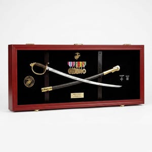 May include: A display case with a red frame, showcasing a saber, medals, and insignia. The saber has a gold handle and a silver blade. The case also features a black interior, a gold emblem, and a small plaque.