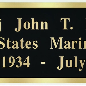 May include: A black plaque with gold lettering and a gold border. The text reads: Sgt Maj John T. Bradsher, United States Marine Corps, May 19, 1934 - July 2, 2003.