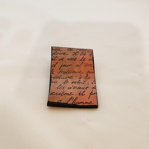 May include: A close-up of a small, rectangular piece of paper with handwritten text in black ink on a light brown background. The text reads: "nde de men, gloire. H là il a créé le u od four il cre le troisième fee reduire à la a le soleil, la les oiseaux do seième il pr a l'homme"