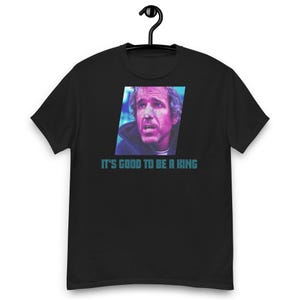 Peut inclure: T-shirt noir avec le texte "IT'S GOOD TO BE A KING" en bleu clair. Le t-shirt présente un graphisme du visage d'un homme dans un schéma de couleurs bleu et rose. Le t-shirt est suspendu à un cintre noir.