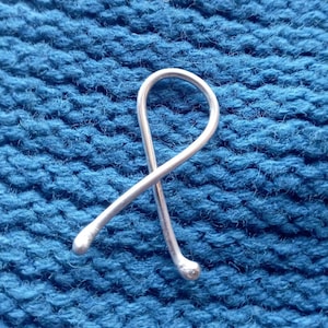 Puede incluir: Un marcador de punto de tejido de color plateado en forma de ocho curvado. El marcador se encuentra sobre un fondo de tejido azul texturizado.