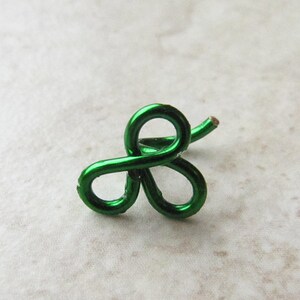 Pode incluir: Um amuleto de trevo de arame verde com três laços. O amuleto é feito de arame e tem um acabamento brilhante.