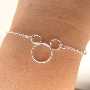 Könnte beinhalten: Ein silbernes Armband mit einem Mickey Mouse-inspirierten Design. Das Armband hat eine feine Kette und einen Anhänger mit einem großen Kreis und zwei kleineren Kreisen, die die ikonische Silhouette bilden. Das Armband wird am Handgelenk getragen.