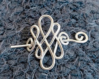 May include: Silver wire hair clip with a decorative knot design. The clip is on a gray fuzzy surface.