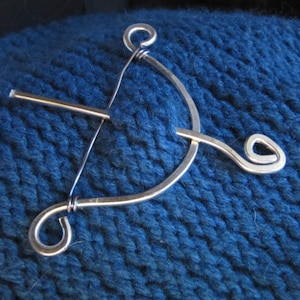 Könnte beinhalten: Eine silberne Schalstecknadel in Form von Pfeil und Bogen. Die Nadel liegt auf einem blauen Strickstoff.