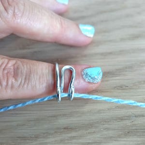 Op de afbeelding: Een zilveren ring met een lus aan de bovenkant, gedragen op een vinger met lichtblauwe nagellak. Een lichtblauwe draad is door de lus geregen.