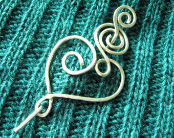 May include: Silver wire heart-shaped shawl pin with a decorative swirl design. The pin is on a teal knitted fabric.