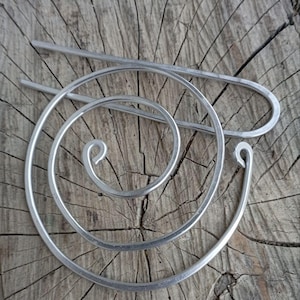 Op de afbeelding: Een close-up van drie zilveren haaraccessoires. De accessoires omvatten een lange, rechte speld, een spiraalvormige haarspeld en een gebogen haarspeld. De items worden weergegeven op een verweerd, houten oppervlak.