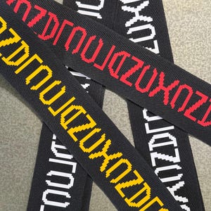 May include: Three black woven fabric straps featuring a repeating pixelated text pattern. One strap displays the text in red, another in yellow, and the third in white. The text appears in a stylized, blocky font with a distinct digital aesthetic.