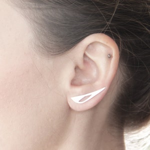 May include: A close-up of a person's ear with a silver ear cuff. The ear cuff is a thin, curved piece of metal that is designed to sit on the upper part of the ear.