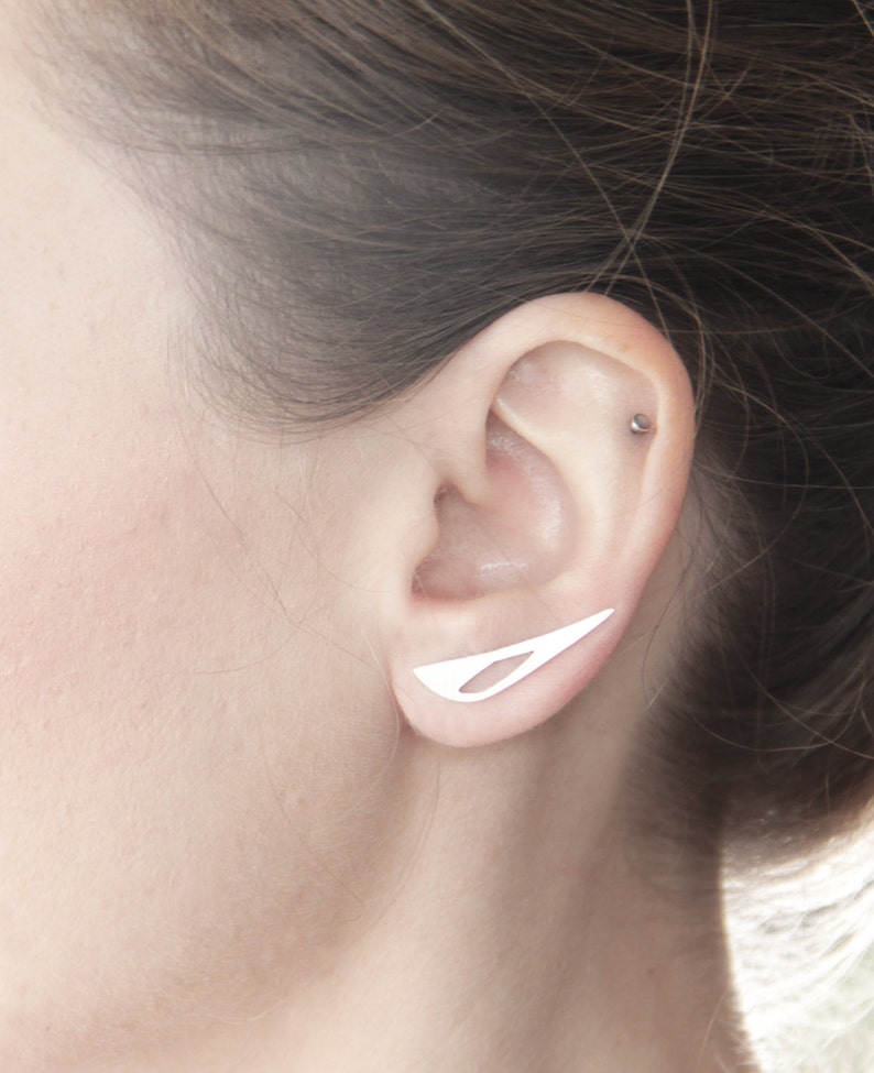 May include: A close-up of a person's ear with a silver ear cuff. The ear cuff is a thin, curved piece of metal that is designed to sit on the upper part of the ear.