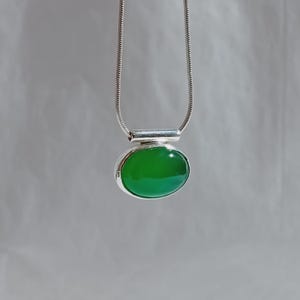 Può includere: Una collana d'argento con un ciondolo ovale verde brillante. Il ciondolo è incastonato in argento lucido e appeso a una sottile catena d'argento. La pietra verde brillante contrasta con l'argento, creando un gioiello suggestivo.