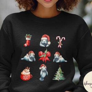May include: Black sweatshirt featuring a festive design with watercolor illustrations. The design includes a Christmas stocking, candy canes, a red bow, a Christmas tree, and manatees wearing Santa hats. The sweatshirt also has a red mug with whipped cream and a logo.