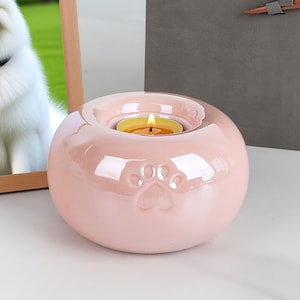 May include: A pink ceramic candle holder with paw print details, holding a lit tea light. A framed photo of a white dog is to the left, and a gray box is in the background.