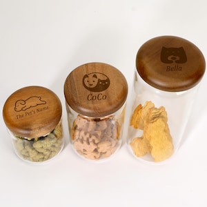 May include: Three clear glass pet treat jars with wooden lids. The lids are engraved with pet-themed designs and names: "The Pet's Name", "CoCo", and "Bella". The jars contain various pet treats in shades of green, brown, and yellow.