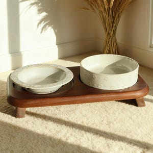 Puede incluir: Un soporte de madera oscura para cuencos para mascotas con dos cuencos de cerámica. Un cuenco tiene un patrón floral, el otro huellas de patas. El soporte es de color marrón oscuro y tiene un diseño curvo.