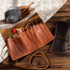 May include: A tan leather crochet hook case, open to reveal hooks and scissors. A ball of grey yarn and a small basket with wooden crochet hooks are in the background. A rolled-up dark brown leather case is also present.