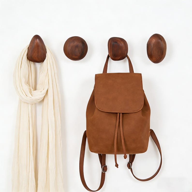 May include: Four wooden wall hooks in varying organic shapes are mounted on a white wall. A light brown leather backpack hangs from one hook, and a cream-colored scarf is draped over another. The hooks are a dark, warm brown.