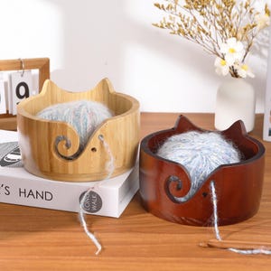May include: Two wooden yarn bowls, one light-colored and one dark-colored, each holding a ball of blue and white yarn. The bowls have a cat-ear design and a swirl-shaped yarn guide. A crochet hook is visible.