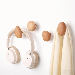 May include: Wooden wall hooks in various shapes and sizes. One hook holds a pair of cream-colored headphones, and another holds a cream-colored scarf. The hooks are made of light-colored wood and are mounted on a white wall.