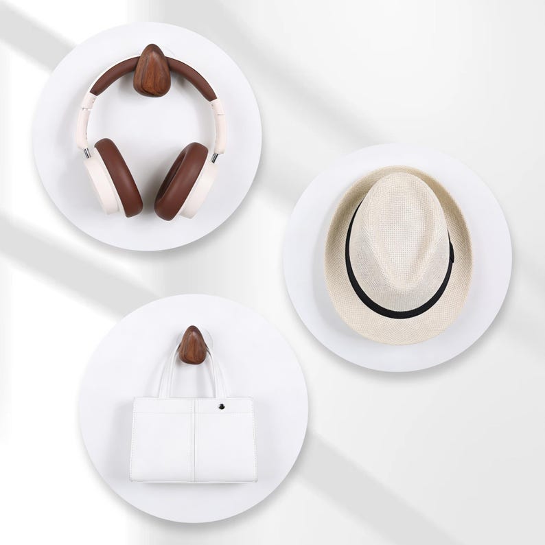 May include: Three white circular wall hooks with wooden accents. One hook holds brown and white headphones, another a straw fedora with a black band, and the third a white handbag. The items are displayed against a white background.