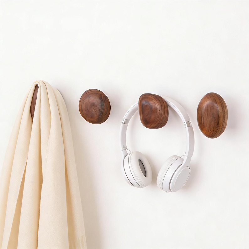 May include: Three wooden wall hooks in a natural wood finish are mounted on a white wall. A white scarf hangs on the first hook. A pair of white headphones hangs on the center hook. The hooks are oval and rounded.