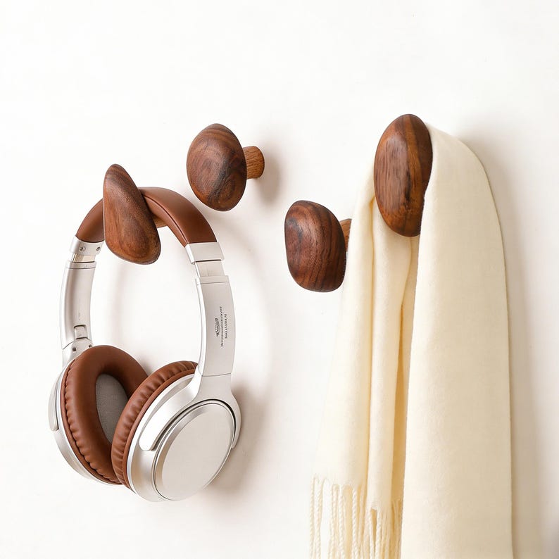May include: Three wooden wall hooks in a natural finish. One hook holds a pair of silver and brown headphones. Another hook holds a cream-colored scarf. The hooks are mounted on a white wall.