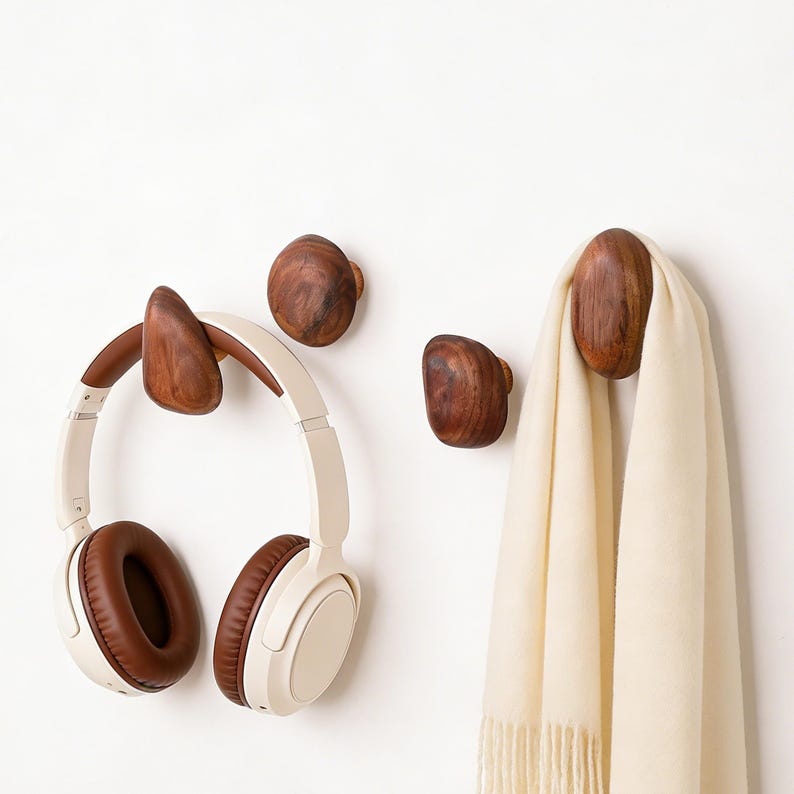 May include: Wooden wall hooks in various shapes and sizes. One hook holds a pair of white and brown headphones. Another hook holds a cream-colored scarf. The hooks are made of dark wood and are mounted on a white wall.