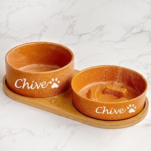 May include: Two orange ceramic pet bowls with the name "Chive" and a paw print on each bowl. The bowls are on a light brown bamboo tray.