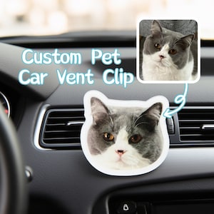 May include: A custom car vent clip shaped like a cat's face, in shades of grey and white. The clip is attached to a car vent. A smaller image of the cat is in the top right. The text on the image reads "Custom Pet Car Vent Clip."