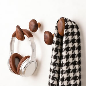 May include: Wooden wall hooks in various shapes and sizes. One hook holds a pair of silver and brown headphones. Another hook holds a black and white houndstooth scarf. The hooks are made of dark wood and are mounted on a white wall.