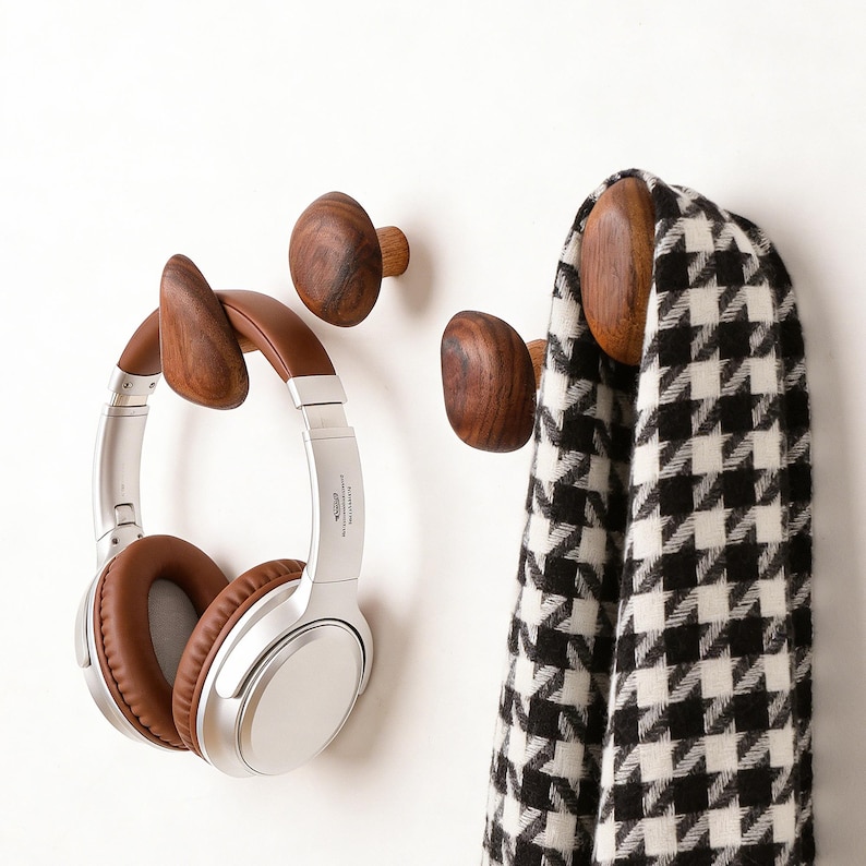 May include: Wooden wall hooks in various shapes and sizes. One hook holds a pair of silver and brown headphones. Another hook holds a black and white houndstooth scarf. The hooks are made of dark wood and are mounted on a white wall.