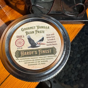 May include: A silver lidded jar with a white label that reads "Gourmet Vanilla Bean Paste, Grade A, 4 fl oz, Made with White Rum & Grade A Madagascar Vanilla Beans, Hardy's Finest. This homemade product was not produced in a licenced or regulated facility. 337.739.5300 117 Ok Avenue, Harahan, LA 70123". The label also features a cartoon image of a bird.