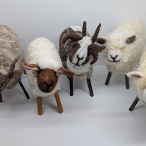 May include: A collection of five needle-felted animal figurines standing on thin wooden legs. The group includes three sheep with cream and white wool, one goat with brown and white markings and horns, and one sheep with grey wool and horns.