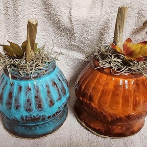 Könnte beinhalten: Zwei Keramik-Kürbisse, die für den Herbst dekoriert sind. Einer ist in blaugrün gehalten, der andere in einem verbrannten Orange. Beide haben einen rustikalen, abgenutzten Look.
