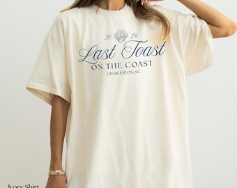 May include: Ivory-colored t-shirt with the text "Last Toast On The Coast Charleston, SC" in navy blue script. The shirt has a relaxed fit and short sleeves. The text includes a seashell graphic and the numbers "20 2x".