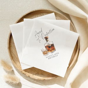 May include: White paper napkins with a decorative design featuring a whiskey bottle and glass, with the text "Aged to Perfection" and "Scott's 40th Birthday". The napkins are arranged on a wooden plate.