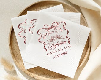May include: Three white square paper napkins featuring a dark red printed design of a decorative ribbon bow framing two wine glasses. The text reads AGED TO Perfection, HANNAH MAY, and 3.12.1986 in a mix of serif and script fonts.
