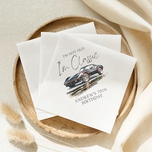 Può includere: Una pila di tovaglioli di carta bianchi con l'illustrazione di un'auto sportiva vintage nera. Il testo recita I'M NOT OLD I'M CLASSIC e ANDREW'S 70TH BIRTHDAY in caratteri neri. I tovaglioli hanno un bordo testurizzato e sono quadrati.