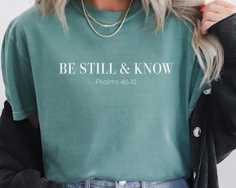 May include: Teal-colored t-shirt with the text "BE STILL & KNOW" and "Psalms 46:10" in white. The shirt is paired with blue jeans and a black cardigan. A gold necklace is visible.