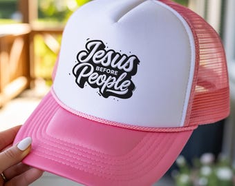 May include: A pink and white trucker hat with the text "Jesus Before People" in black script. The hat has a pink brim and a white front panel with a mesh back. The hat is being held up by a person.