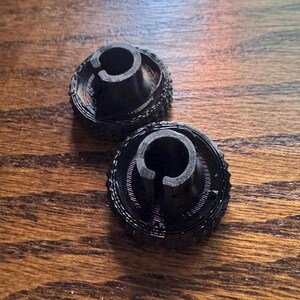 May include: Two black, 3D-printed plastic objects. Each has a circular base with a textured edge and a central, cylindrical post with a curved cutout. The objects are on a wooden surface.