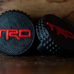 Op de afbeelding: Twee zwarte kunststof wieldoppen met een rood "TRD" logo. De doppen hebben een getextureerd oppervlak en een verhoogde rand.